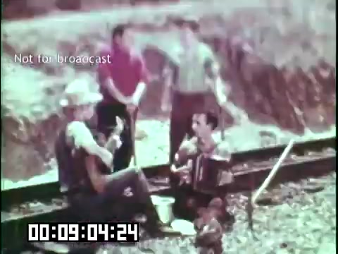 The still from 'Immigrant Heritage' features a group of individuals gathered on a railway line, engaged in a musical session. One person is playing an accordion, while others appear to be enjoying the performance. The scene captures a sense of camaraderie and cultural expression, reflective of the immigrant experience, with a soft color palette typical of 16mm film. The timestamp and 'Not for broadcast' overlay indicate this is a clipped segment meant for educational purposes.