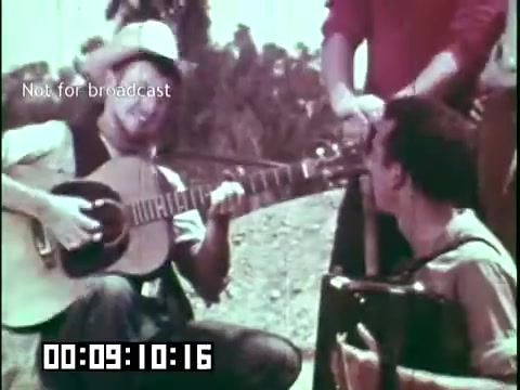 The still from 'Immigrant Heritage' features two musicians in a lively outdoor setting. One musician plays an acoustic guitar while sitting, sporting a wide-brimmed hat, while the other plays an accordion and looks on appreciatively. The scene captures a moment of camaraderie and cultural expression, highlighting the Irish Rovers' connection to music and heritage. The film likely focuses on themes of immigration and cultural identity through traditional music.