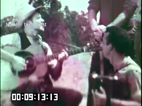 The still from 'Immigrant Heritage' features two musicians performing in a natural outdoor setting. One person is strumming a guitar while the other listens intently, showcasing a moment of musical collaboration. The vibrant colors indicate an artistic approach typical of the era, and the scene captures the cultural essence of the Irish Rovers, emphasizing themes of heritage and community through music. The timestamp suggests a segment of the film focused on performance.