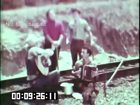 The still from 'Immigrant Heritage' featuring the Irish Rovers shows a group of four individuals engaged in a musical performance. One person plays a guitar while another plays an accordion, suggesting a lively folk music session. They appear to be seated outdoors, potentially on a train track, with a rural backdrop. The atmosphere seems joyful, indicative of cultural celebration and community spirit.