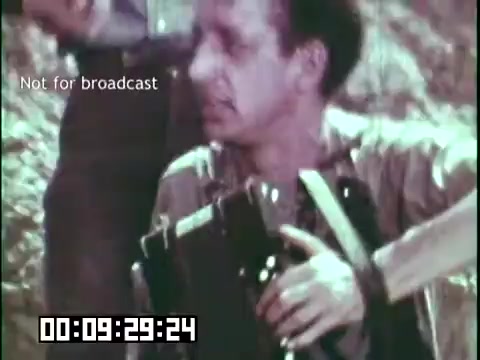 The still from 'Immigrant Heritage' features a man holding an instrument, likely part of a musical performance. He appears engaged and animated, suggesting a lively moment in the film. The background hints at an outdoor setting, adding to the cultural context of the Irish Rovers and their connection to immigrant heritage. The timecode indicates that this scene occurs early in the film.