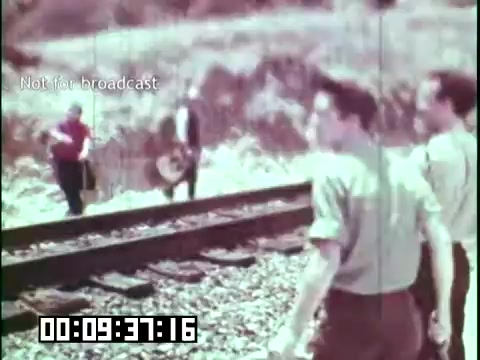 The still from the educational film 'Immigrant Heritage' featuring the Irish Rovers shows a group of individuals near railway tracks. Two figures are walking away from the camera, with one holding a guitar, while a third person is visible in the background engaging with another individual. The scene captures a nostalgic moment, likely depicting themes of cultural heritage and community connection through music. The color tone appears faded, characteristic of 16mm film, adding to the vintage feel of the image.