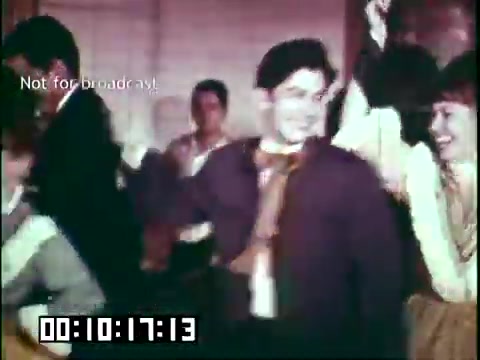 The still is a vibrant moment from the educational film 'Immigrant Heritage,' featuring a lively scene likely depicting a cultural celebration. People are engaged in traditional dancing, dressed in festive attire, with expressions of joy and enthusiasm. The setting appears to be a gathering, emphasizing community and cultural heritage. The timecode at the bottom suggests this scene occurs shortly into the film, and the watermark indicates it is not for broadcast.