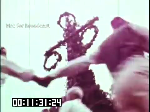 The still from 'Immigrant Heritage' featuring the Irish Rovers captures a lively scene of celebration. In the image, individuals are holding hands, possibly dancing or participating in a folk tradition. The background features a decorative structure, likely symbolizing heritage or community. The visual conveys a sense of joy and cultural connection, reflecting the themes of the film about immigrants and their traditions. The timestamp suggests it is part of a sequence showcasing cultural practices.