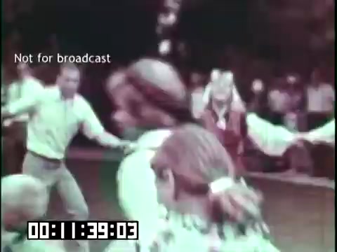 The still from 'Immigrant Heritage,' featuring the Irish Rovers, depicts a lively scene with a group of people dancing in a circle outdoors. The image captures the spirit of community and celebration, reflecting cultural heritage. People are dressed in casual attire, and the atmosphere appears joyful and festive, highlighting traditional dance as a key element of the immigrant experience.