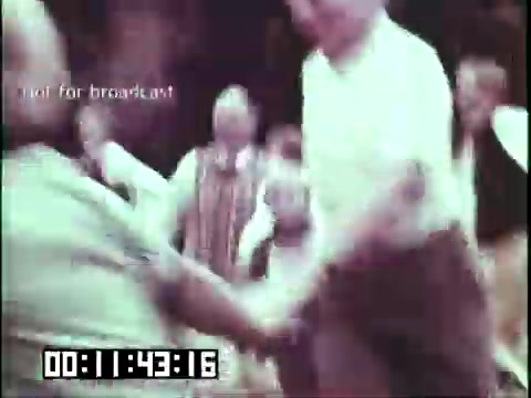 The 16mm still from 'Immigrant Heritage' features a lively scene with people dancing in a festive setting. The focus is on individuals engaged in traditional dance, showcasing cultural heritage. The atmosphere appears joyous, with colorful clothing suggesting a celebration of Irish culture. The timecode indicates a specific point in the film, hinting at active engagement in the narrative of immigrant experience.