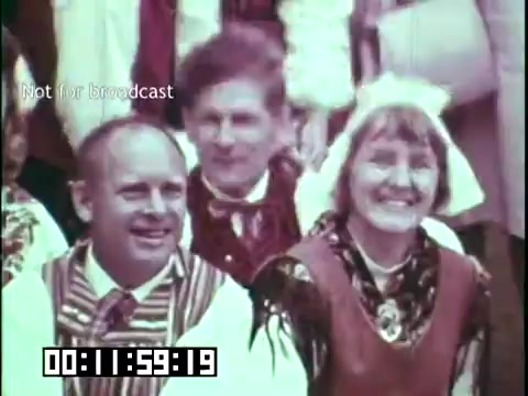 The still from 'Immigrant Heritage' features a group of individuals wearing traditional cultural attire, suggesting a celebration of heritage. Two prominently smiling figures in the foreground appear to be enjoying a moment of community or performance. The colorful clothing reflects the rich traditions associated with immigrant cultures, likely emphasizing the film's themes of identity and heritage. The timestamp indicates a specific moment within the film, which appears to capture joyful participation in cultural expression.