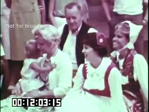 The still from 'Immigrant Heritage' features a diverse group of individuals gathered in a lively outdoor setting. In the foreground, a woman holds a child, while others, dressed in traditional or cultural attire, engage joyfully. The backdrop appears to be a festive environment, suggesting themes of community, heritage, and celebration. The scene emphasizes cultural identity and connection among different generations.