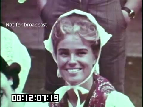 The still from 'Immigrant Heritage' features a smiling young woman wearing traditional attire, complete with a white bonnet and colorful dress. Her expression conveys a sense of joy and cultural pride. The background appears to be outdoor, possibly during a festival or cultural event, reflecting the themes of heritage celebrated in the film. A timestamp at the bottom indicates the specific moment in the film. The label 'Not for broadcast' suggests it's for private viewing or archival purposes.