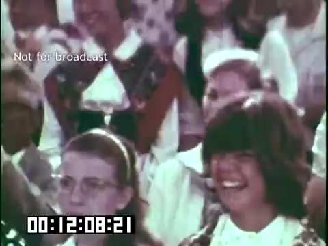 The still from 'Immigrant Heritage' features a lively audience engaged in a performance, likely by the Irish Rovers. The crowd appears diverse, with individuals wearing various traditional outfits. Notable in the image are smiles and laughter, suggesting an atmosphere of enjoyment and appreciation for cultural heritage. The timecode indicates it is a segment from the film, emphasizing its educational purpose.