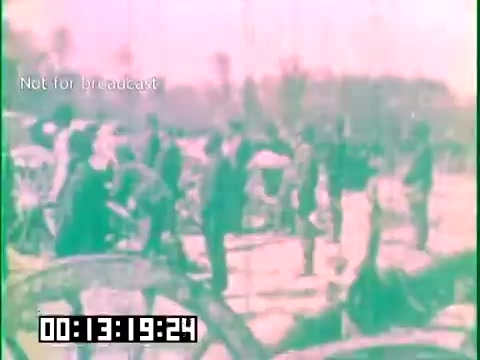 The 16mm still from 'Immigrant Heritage' features a group of people, likely immigrants, engaged in various activities outdoors. The scene appears historical, with a soft color palette suggesting an older film style. There are trees in the background, indicating a natural setting, and the individuals are dressed in period attire. The timestamp and 'Not for broadcast' notice at the bottom suggest this is a segment from the film, focusing on the heritage and experiences of immigrant communities, possibly highlighting their labor and resilience.