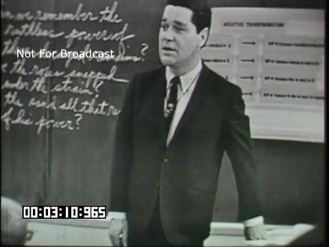 The still features a male instructor in a suit standing in front of a classroom blackboard. He is engaged in a lecture, addressing students off-camera. The blackboard contains notes and questions related to linguistics, reinforcing the educational theme of the film. The visual style suggests a mid-20th-century educational context, with an emphasis on traditional teaching methods.