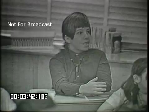 The still features a young student in a classroom setting, engaged in discussion. He has a thoughtful expression and is sitting at a desk with his arm resting on it. The classroom appears traditional, with wooden desks and a chalkboard in the background. The film is marked 'Not For Broadcast' and displays a timestamp at the bottom. It captures a moment intended to illustrate a teaching scenario related to linguistics for a sixth-grade audience.