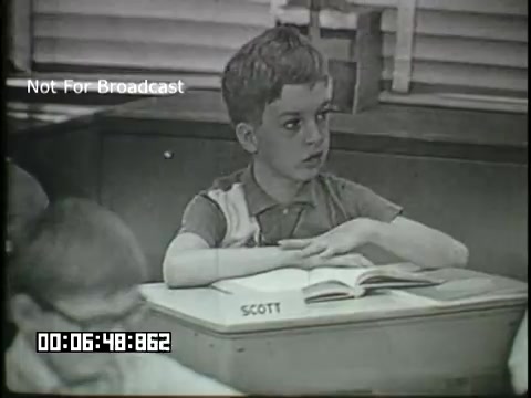 The still from the 'Sixth Grade Linguistics' educational film features a young boy named Scott sitting at a classroom desk. He appears attentive, with his hands crossed in front of him. A book is open on his desk, and there are blurred classmates in the foreground. The classroom has wooden elements and blinds in the background, suggesting a typical mid-20th century educational setting. The image conveys a focused learning environment.