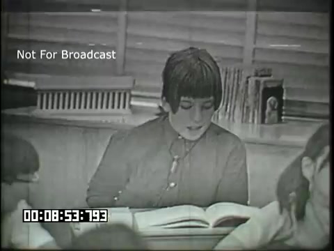 The still from 'Sixth Grade Linguistics' depicts a classroom scene where a student, wearing a button-up shirt, is reading from a book. The focus is on the student, who appears engaged and expressive, while a few classmates are seen in the background, some of whom are attentive to the reading. The environment suggests a typical educational setting, reinforced by classroom elements like windows and bookshelves. The film uses a vintage aesthetic characteristic of educational media from that era.