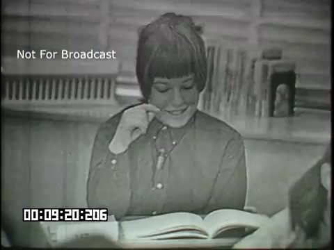 The still features a smiling young girl with short hair, sitting at a desk and engaging with a large book. She appears to be participating in an educational setting, likely discussing or learning about linguistics. The classroom background suggests a focus on interactive learning, with resources visible behind her. The scene captures a moment of engagement and curiosity in a sixth-grade context.