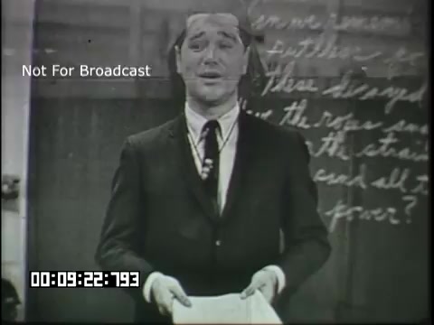 The still from the 'Sixth Grade Linguistics' educational film features a man dressed in a suit, standing in front of a blackboard with handwritten notes. He appears to be mid-sentence, possibly engaging with an audience or students. The setting suggests a classroom environment, highlighting a topic related to linguistics. The timestamp indicates that it is part of a longer presentation. The image is marked 'Not For Broadcast,' implying it may be for educational use only.