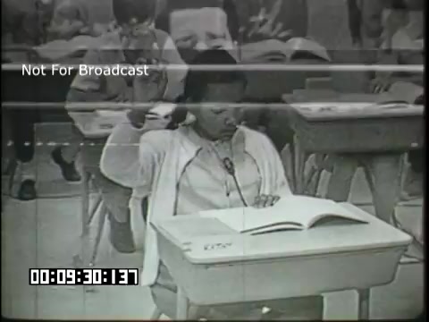The still from the 'Sixth Grade Linguistics' educational film features a classroom scene where a student, seated at a desk, has raised their hand, presumably to answer a question or participate in a discussion. The student is focused, and a book is open on the desk, indicating an active learning environment. Other students in the background appear engaged as well. The image captures a moment of interaction typically found in a sixth-grade classroom setting.