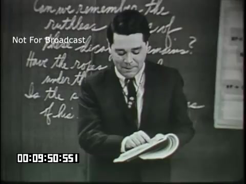 The still features a male educator standing in front of a blackboard filled with handwritten notes. He appears to be engaged in teaching, holding an open book in one hand while referencing the content. The educator is dressed in a suit and tie, suggesting a formal classroom setting. The blackboard contains phrases likely related to linguistic concepts, indicating the educational theme of the film directed towards sixth-grade students. A timestamp at the bottom suggests it is a clip from a longer presentation.