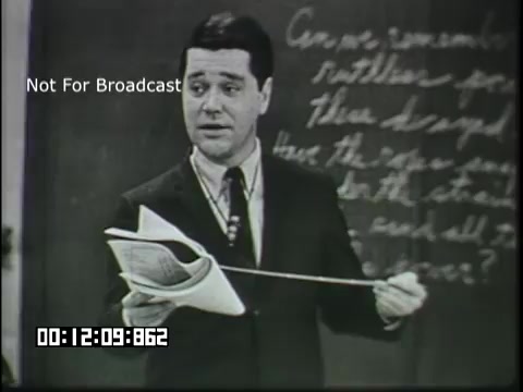 The still features a male teacher standing in front of a blackboard filled with writing. He is dressed in a formal outfit with a tie, holding a sheet of paper and possibly pointing to key concepts as he addresses students. The overall setting suggests a classroom environment, aimed at educating sixth graders about linguistics. The tone appears serious yet engaging, typical of educational films from that era.