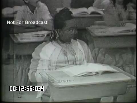 The still features a classroom setting from the 'Sixth Grade Linguistics' educational film. A student is sitting at a desk with an open book, appearing engaged in learning. Other students are visible in the background, also seated at their desks. The monochrome film captures the atmosphere of a 1960s educational environment. 'Not For Broadcast' is overlaid on the image, with a timestamp indicating the specific moment in the film.