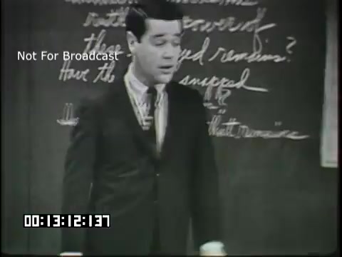The still shows a young man in a suit standing in front of a chalkboard covered with handwritten notes. He appears to be engaged in teaching, possibly discussing linguistic concepts relevant to sixth graders. The scene captures an educational moment, emphasizing the interaction between teacher and students. The time code at the bottom indicates the film's progression.