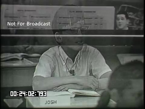 The still features a classroom setting from the 'Sixth Grade Linguistics' educational film. A boy named Josh is seated at his desk, focused on the lesson. He is wearing a light-colored shirt and glasses. In the background, various classroom elements can be seen, including charts or posters related to the linguistic theme of the film. The ambiance suggests a typical learning environment aimed at middle school students.
