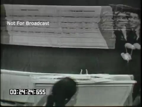 The still from the 'Sixth Grade Linguistics' educational film depicts a classroom setting with a focus on a teacher and students engaged in a lesson. It features a white board or a large paper surface that likely displays linguistic concepts or writing exercises. The overall atmosphere is educational, with an emphasis on interaction and learning. The timestamp indicates it's part of a segment, emphasizing a structured format typical of educational films from that era.