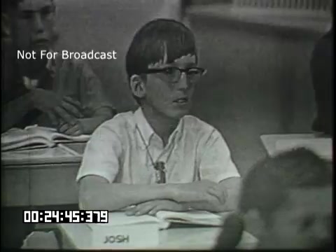 The still from the 'Sixth Grade Linguistics' educational film features a classroom setting. A young boy with glasses and short hair appears to be attentive, sitting at a desk with his hands on the table. The atmosphere suggests a focus on learning, with other students visible in the background, creating an engaging educational environment. The image captures a moment in a learning context related to linguistics.