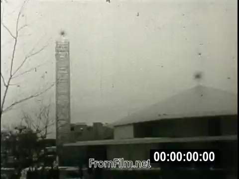 The still features a scene from the 1964 New York World's Fair, highlighting the pavilion dedicated to evangelist Billy Graham. The image captures the pavilion structure, which is likely characterized by its unique architectural elements, alongside a tall monument or tower in the background. It's a moment in time, emphasizing Graham's influential role and message during a period marked by growing materialism and secularism. The context reflects the significance of spiritual engagement in the larger framework of the fair, where diverse cultural and technological innovations were showcased.