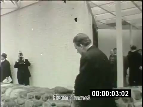 The still captures a moment from a 16mm newsreel featuring Billy Graham at the 1964 New York World's Fair. Graham is seen walking near a stone structure, with a few individuals in suits positioned nearby. One person appears to be filming him. The scene embodies the atmosphere of a significant cultural event where Graham addresses themes of spirituality in a materialistic era. His presence at the fair highlights the intersection of faith and public life during that period.