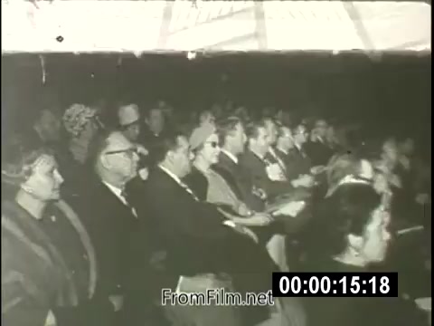 The still captures a moment from a 1964 newsreel featuring Billy Graham speaking at the New York World's Fair Pavilion. In the image, an audience is gathered, attentively listening to him. The setting appears formal, with attendees dressed in mid-20th century attire. Graham's speech emphasizes the significance of faith in a materialistic society, reflecting the cultural and religious dynamics of the era. The atmosphere suggests engagement, highlighting the event's importance in promoting spiritual discussions amidst the fair's attractions.