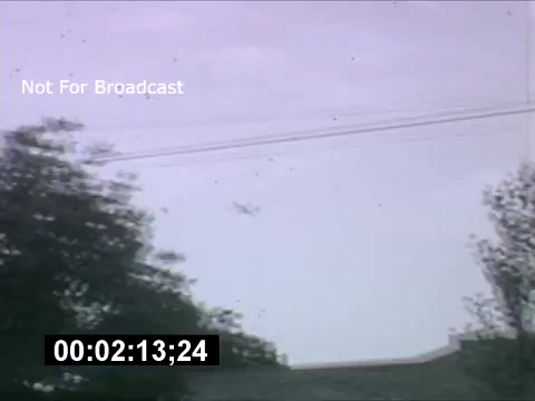 The still depicts a cloudy sky with trees in the foreground. A B-36 bomber can be seen flying above, likely high up and perhaps slightly obscured. The image indicates a frame from 1950s home movies, with a timestamp visible at the bottom. The colors are muted, typical of that era’s film quality.