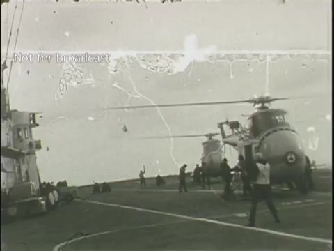 The still appears to show a scene from a naval ship or aircraft carrier, featuring two helicopters on the deck. In the background, personnel are engaged in activities, potentially preparing for a flight or rescue operation. The footage is in black and white, indicating its age, and displays visual artifacts, such as scratches and grain, typical of 16mm film. The overall context relates to news coverage of emergency situations, possibly including responses to plane crashes.