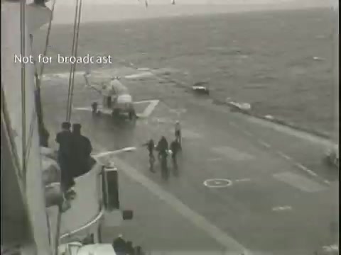 The still shows a scene on a ship's deck, likely an aircraft carrier, where a helicopter is preparing for takeoff or landing. The weather appears overcast, contributing to a somber atmosphere. In the foreground, a group of individuals is present on the deck, possibly crew members or personnel overseeing operations. The surrounding water looks choppy, suggesting rough conditions. The image is marked 'Not for broadcast,' indicating it may be intended for internal use or archival purposes.