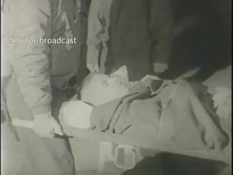 The still depicts a somber scene showing emergency responders carrying an injured individual on a stretcher. The person is wrapped in a blanket, suggesting they may be in critical condition. The black and white footage adds to the gravity of the situation, capturing a moment likely related to a plane crash, reflective of the urgent and distressing nature of the news coverage. The text 'Not for broadcast' indicates this footage is intended for licensing purposes only.