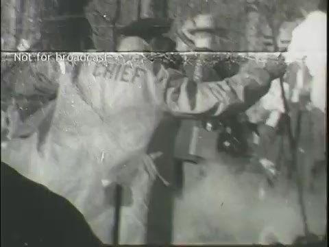 The still captures a chaotic scene from a news report about a plane crash. A figure wearing a jacket labeled 'CHIEF' is seen in the foreground, involved in directing operations. Smoke or debris is present, indicating the aftermath of the incident. Behind the chief, onlookers and emergency personnel are visible, highlighting the urgency and gravity of the situation. The overall atmosphere conveys distress and activity typical of a disaster response.