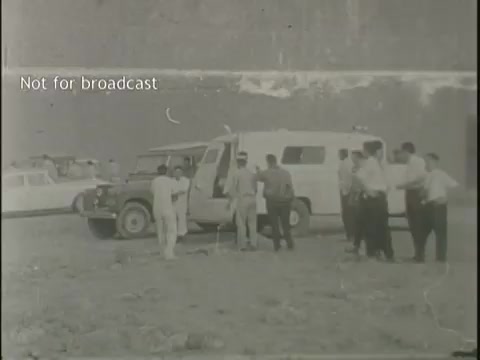 The still features a group of people gathered near an ambulance, amidst a backdrop that suggests an incident site. Several individuals appear to discuss or assess the situation, with vehicles in the background indicating emergency or news response. The monochrome image captures a somber atmosphere, reflective of the serious context of plane crashes and related events. The notation 'Not for broadcast' indicates the footage is restricted from public viewing.