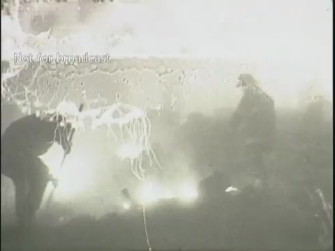 The still depicts a chaotic scene following a plane crash, characterized by a hazy, smoke-filled environment. Firefighters or rescue personnel appear to be actively working among the wreckage, illuminated by flames, while debris is scattered throughout the frame. The atmosphere conveys urgency and the aftermath of a disaster, typical of news footage documenting such tragic events.