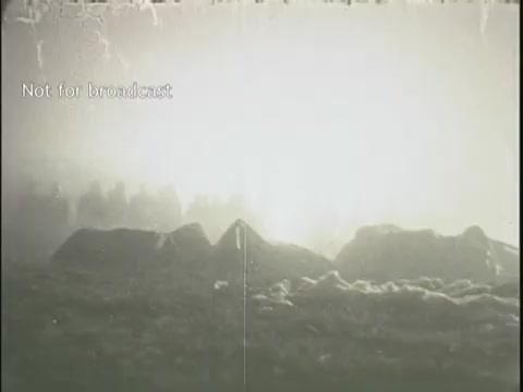 The still shows a hazy, white-lit scene, likely depicting the aftermath of a plane crash. In the background, indistinct figures are visible, possibly rescue personnel or onlookers. The foreground features jagged terrain, which could represent debris or the crash site itself. The image conveys a somber and chaotic atmosphere typical of disaster-related news footage.