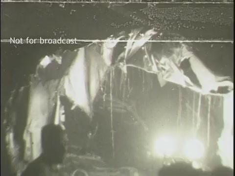 The still captures a chaotic and grim scene from the aftermath of a plane crash. It features twisted metal and debris, suggesting significant destruction. In the foreground, a figure is partially visible, amidst what appear to be emergency lights illuminating the somber surroundings. The overall atmosphere conveys the urgency and seriousness of the situation. The text 'Not for broadcast' indicates that this footage is restricted from public airing.