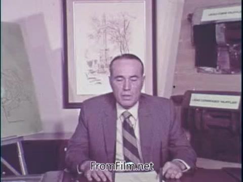 The still from the 'Unleaded Gas Info' film segment features a man seated at a desk, discussing the benefits of lead-free gasoline. He notes that cars using this fuel exhibit significantly improved durability for components like mufflers and spark plugs. Behind him, there are visual elements, including charts or drawings, that support his statements. The overall setting appears to be a formal presentation, likely aimed at educating viewers about the advantages of unleaded gasoline around the time of its introduction in the 1970s.