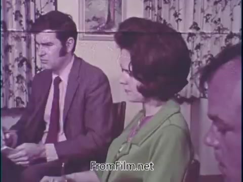 The still features a group of three people engaged in a discussion about unleaded gasoline, likely in a formal or informational setting.   - **Man on the left**: Dressed in a suit and tie, appears to be listening intently, conveying a serious demeanor. - **Woman in the center**: Wearing a green outfit, she seems to be reflecting on the conversation, possibly taking notes or engaged in thought. - **Man on the right**: More casually dressed, he appears to be part of the discussion but less prominent in expression.  The backdrop includes patterned curtains, suggesting a domestic or office environment. The tone of the scene hints at a blend of informative discussion and community concern regarding the introduction of unleaded gasoline.