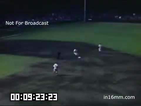 The still features a scene from a Detroit Tigers game at Briggs Stadium, captured between 1948 and 1950. The footage, in Kodachrome, showcases players on the field, likely in the midst of gameplay, with a focus on a grassy area where action is occurring. The low-angle shot highlights the stadium's ambiance, with spectators visible in the background, under the glow of stadium lights. The timestamp indicates it's part of a longer sequence, emphasizing the historical significance of the moment.