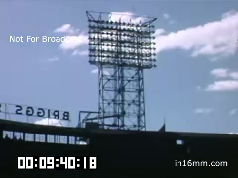 The still shows a view of Briggs Stadium, home of the Detroit Tigers, during the late 1940s. The focus is on a tall lighting tower adorned with numerous lights, set against a bright sky with fluffy clouds. The iconic 'BRIGGS' sign is visible in the foreground, establishing the location. The image captures the nostalgic atmosphere of a baseball game during that era, showcasing the vintage aesthetic of Kodachrome film. The timestamp indicates a segment of this archival footage.