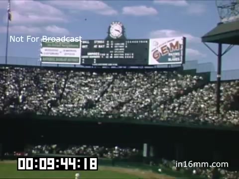 The still features a vibrant scene from a Detroit Tigers game at Briggs Stadium, captured in the late 1940s. The image shows a packed stadium with numerous fans in the stands, under a clear blue sky dotted with clouds. A scoreboard, displaying game details, is prominently visible, along with colorful advertisements. Overall, the shot conveys the lively atmosphere of a baseball game during that era, highlighting the excitement and community spirit of attending a Tigers match.