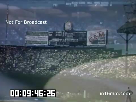 The still captures a vibrant scene from a Detroit Tigers game at Briggs Stadium between 1948 and 1950. It features the scoreboard, which displays information like the game details and perhaps advertisements. The stadium appears lively, with a large crowd visible, and there's a sense of excitement in the air, enhanced by the bright colors typical of Kodachrome footage from that era. The timestamp indicates that this is taken from a specific part of the footage, emphasizing its historical significance.
