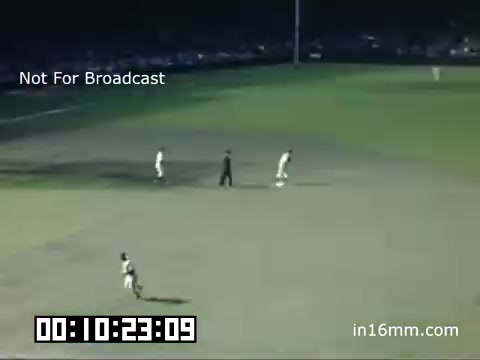 The still shows a scene from a Detroit Tigers game at Briggs Stadium, captured in Kodachrome between 1948 and 1950. In the image, two players are positioned on the field while an umpire stands nearby, suggesting a moment during play. The field is brightly lit, highlighting the grass and players' uniforms, with a crowded spectator area in the background. The timestamp implies this is part of a longer sequence, emphasizing the historical nature of the footage.