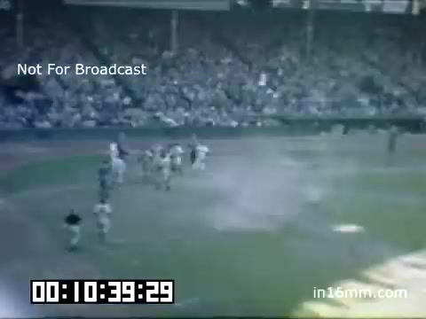 The still captures a scene from a Detroit Tigers game at Briggs Stadium during the late 1940s to 1950. The footage, in Kodachrome, shows a vibrant crowd in the stands, highlighting the excitement of the game. Players are visible on the field, and the atmosphere is lively, with a noticeable sense of engagement from the fans. The timestamp suggests this is part of a longer sequence, emphasizing the historical significance of the moment. The audio track features 'Fuzzball Parade' by Kevin MacLeod, adding a playful backdrop to the visuals.