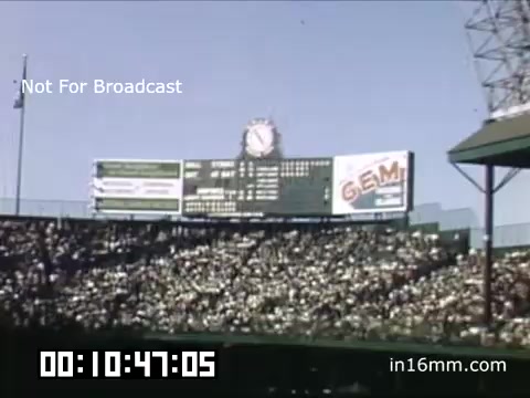 The still depicts a scene from a Detroit Tigers game at Briggs Stadium between 1948 and 1950. The shot features a bustling crowd filling the stands, showcasing the energetic atmosphere of a baseball game. In the background, a scoreboard displays game information, along with advertisements, including one for 'GEM.' The clear blue sky above adds to the overall nostalgic vibe of the Kodachrome footage, highlighting a moment in baseball history. The visual quality reflects the era's film technology, capturing the excitement of the game day experience.